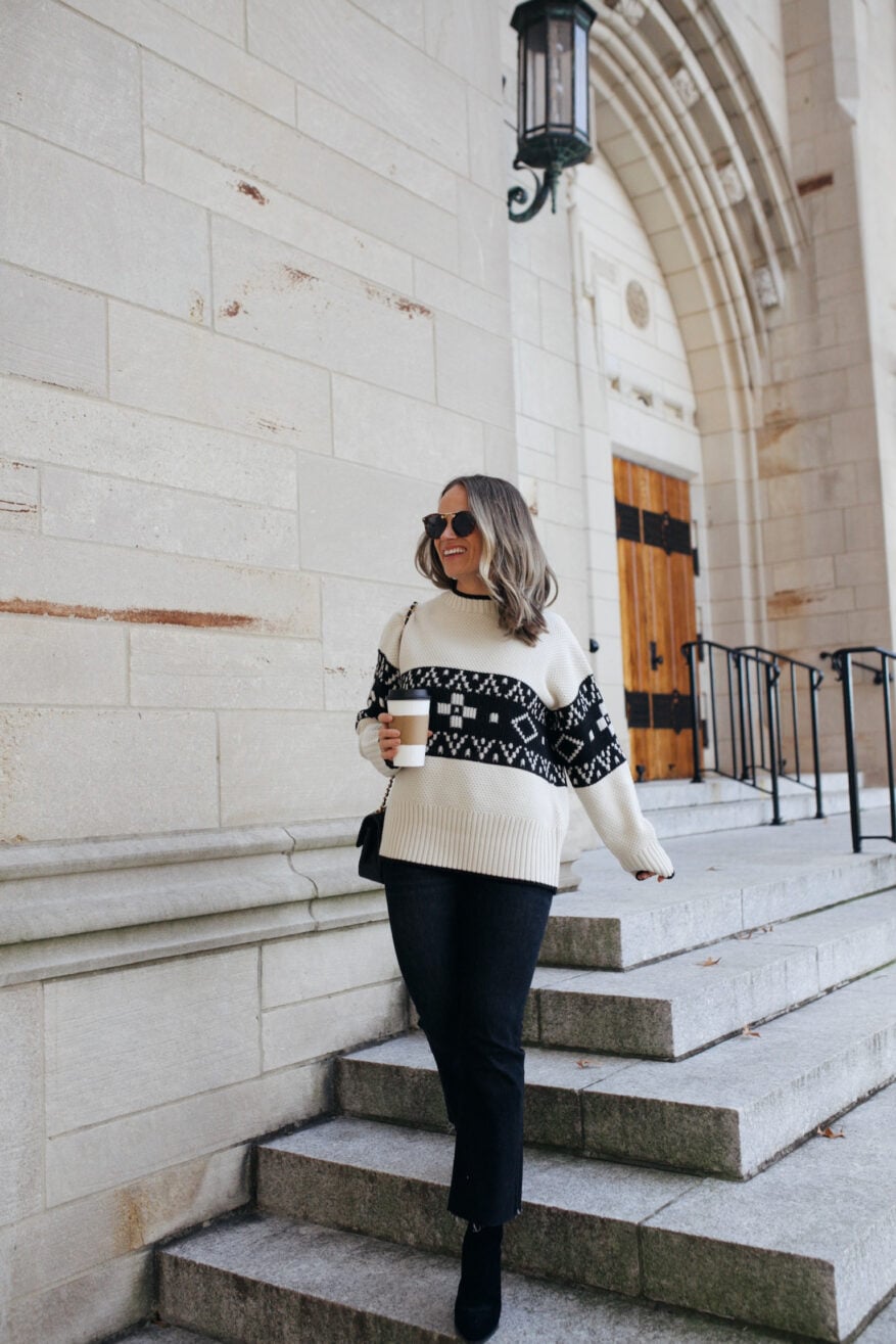 Woman walking down stairs in Christmas Fair Isle Sweater and black jeans with black booties while holding coffee