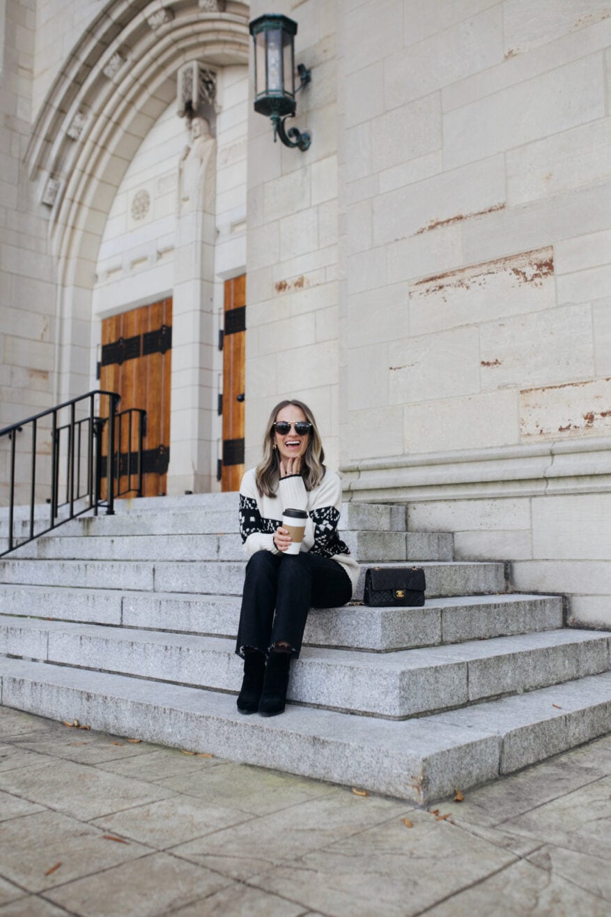 Woman sitting on steps with sunglasses while wearing a black and white Fair Isle sweater with black pants and boots