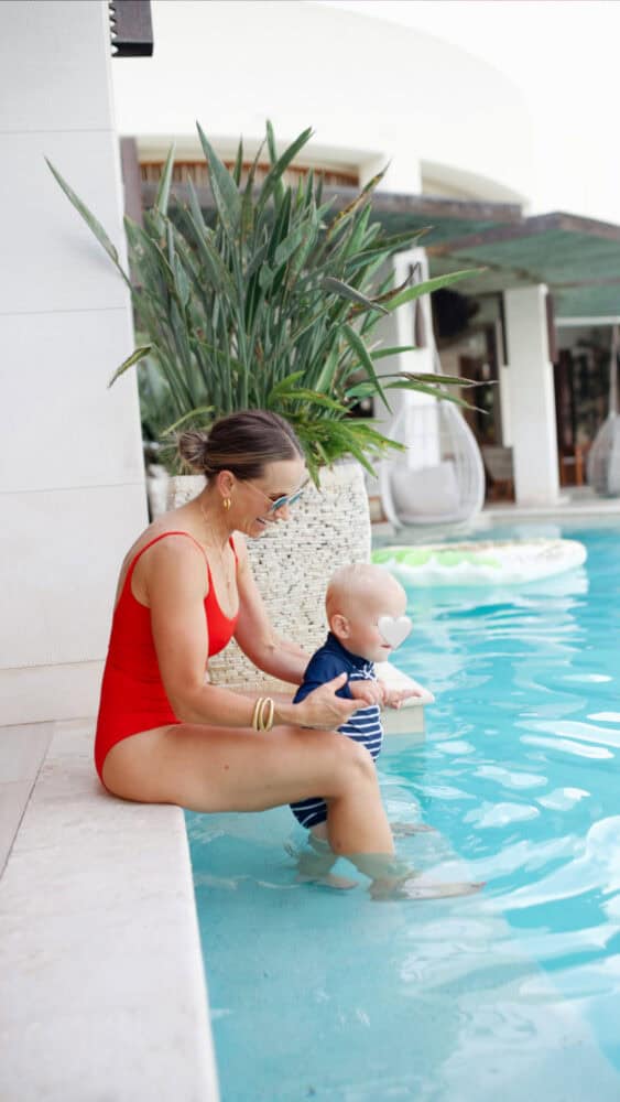 Woman in red bathing suit holding a baby while sitting on side of pool