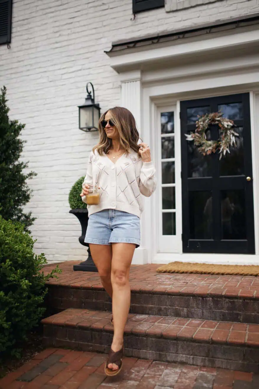 Woman walking down stairs in denim shorts, white sweater, and sandals