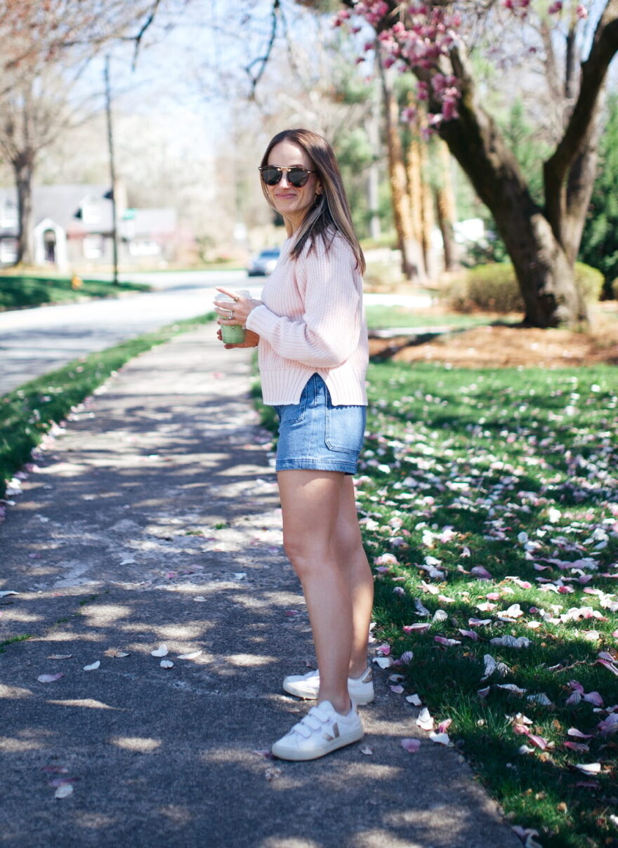 petite woman in white sneakers paired with denim shorts and a long sleeved top under a blooming tree