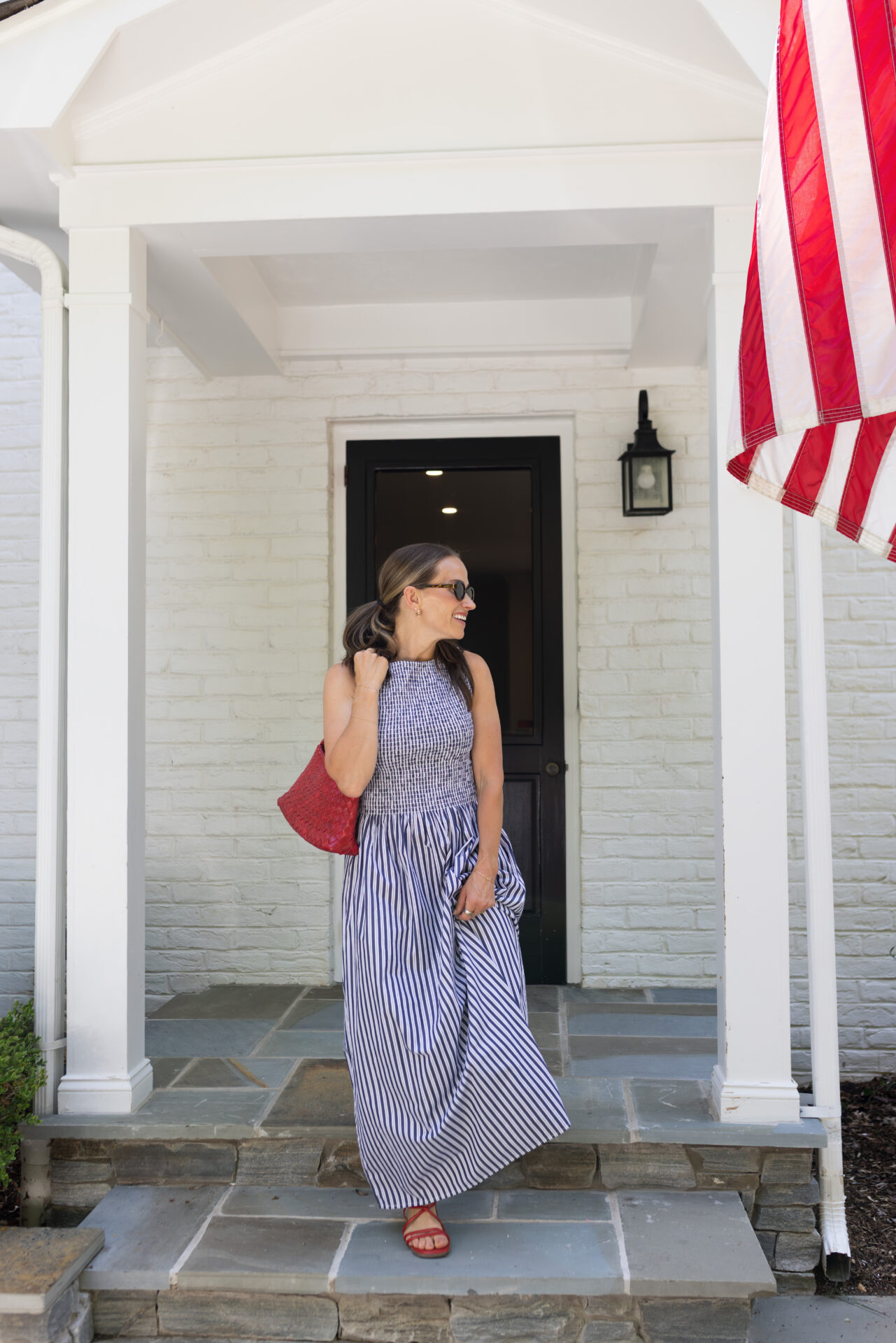 TeriLyn Adams smiling in Hill House Cosima dress navy stripe holding red bag on white brick porch with American flag