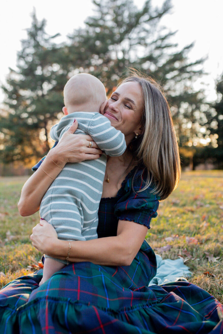 TeriLyn Adams hugging baby while wearing Hill House Louisa dress in navy plaid sitting on grass at sunset