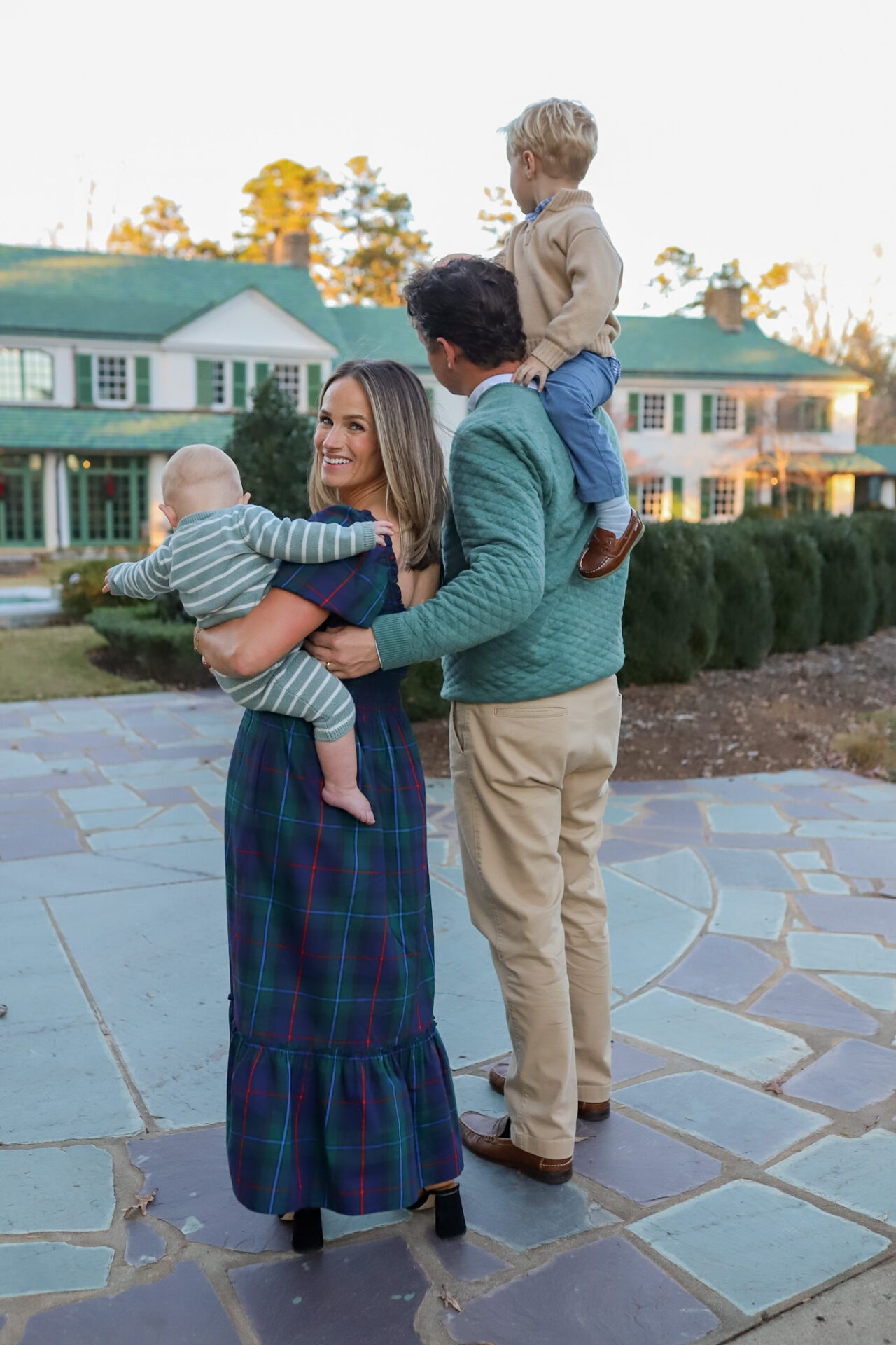 TeriLyn Adams in Hill House Louisa navy plaid dress holding baby while husband carries toddler on shoulders outside estate