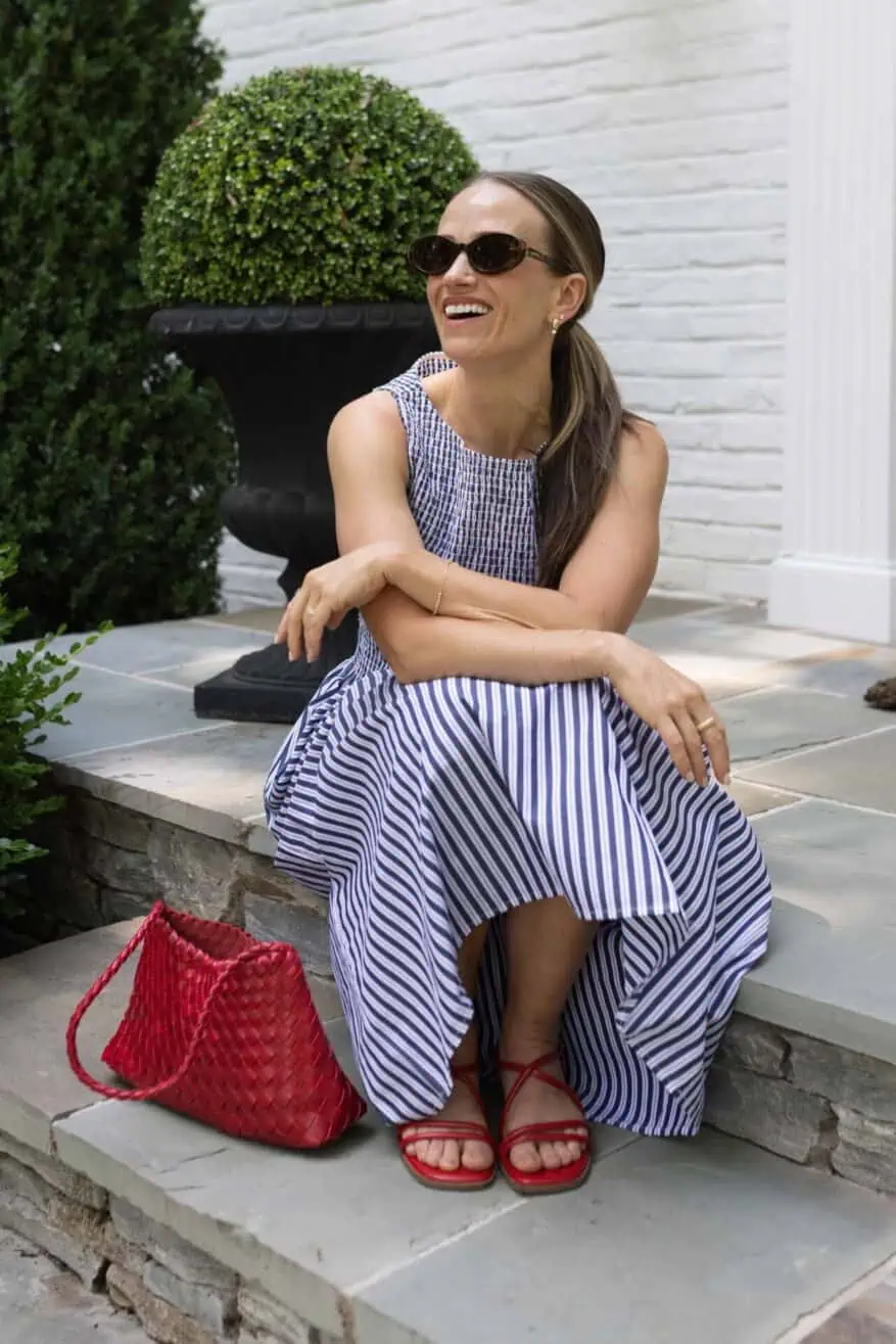 TeriLyn Adams laughing in Hill House Cosima dress navy stripe with red woven bag and red strappy sandals on stone steps