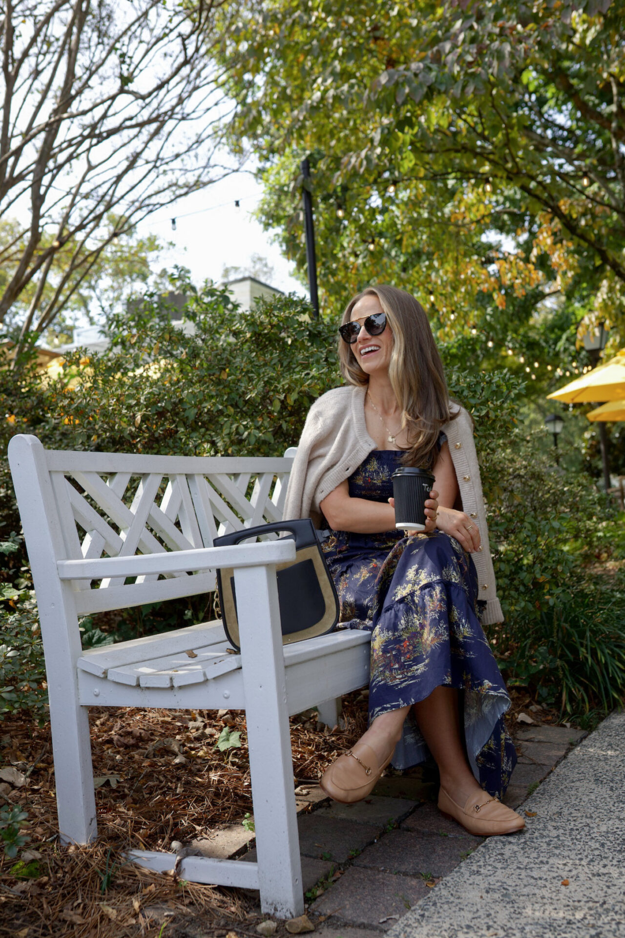 TeriLyn Adams laughing in Hill House Ellie navy floral dress with cream cardigan and tan loafers on white garden bench