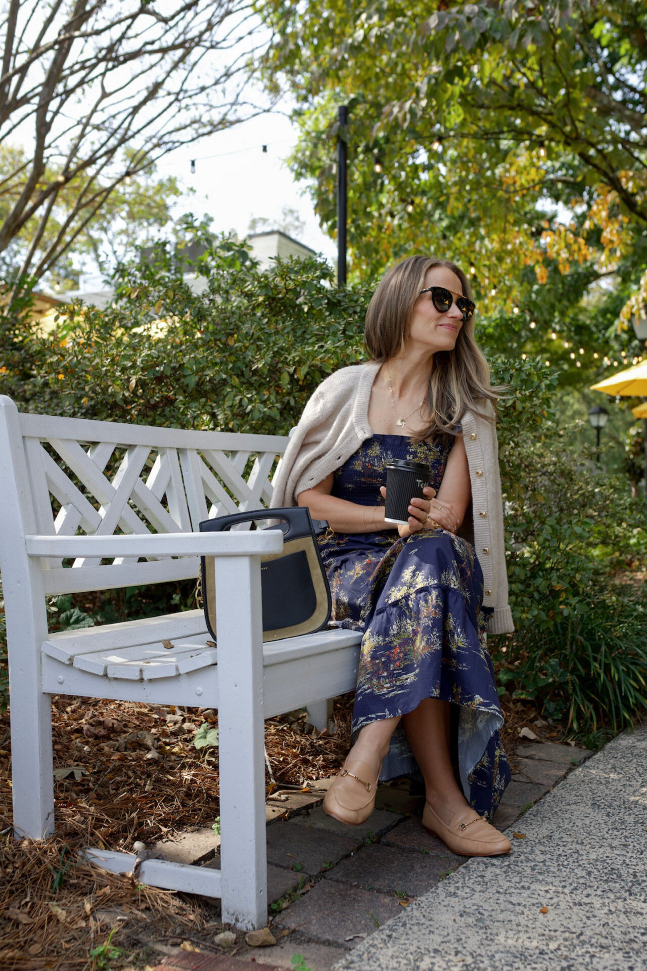 TeriLyn Adams in Hill House Ellie navy floral dress with cream cardigan and tan loafers sitting on white garden bench with coffee