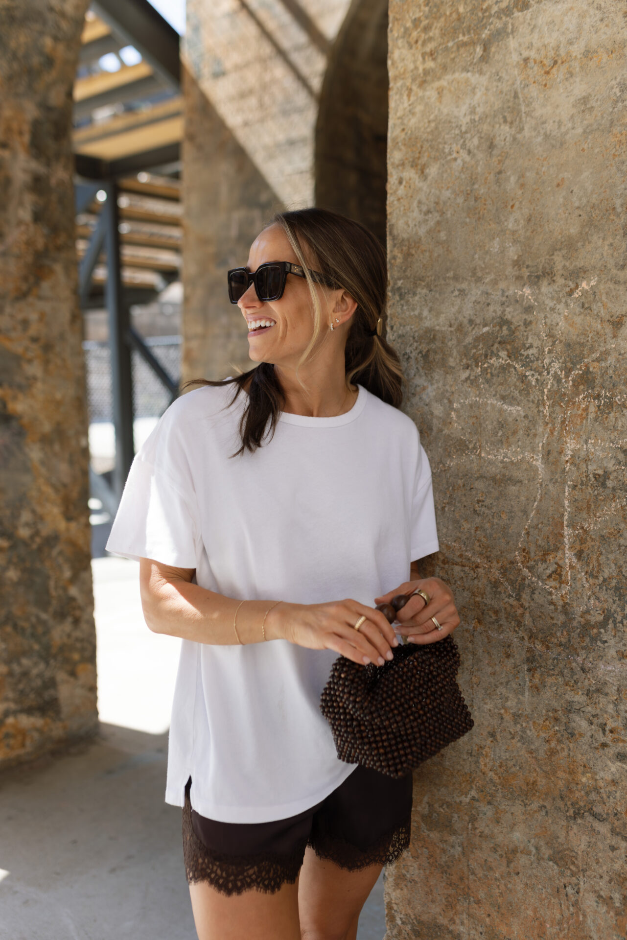 TeriLyn Adams in white tee and lace shorts holding beaded bag wearing Mejuri gold bracelet and stacked rings outdoors