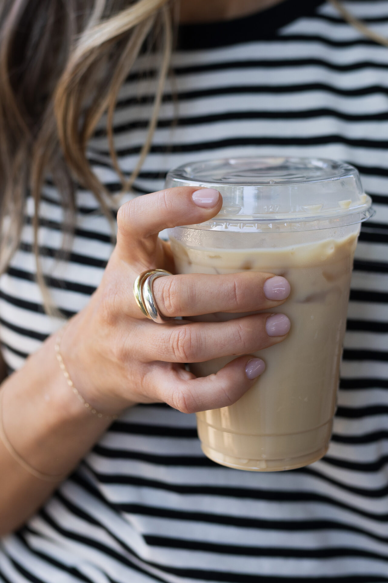 Close-up of hand holding iced coffee wearing Mejuri gold stacked rings and pink manicure in striped shirt