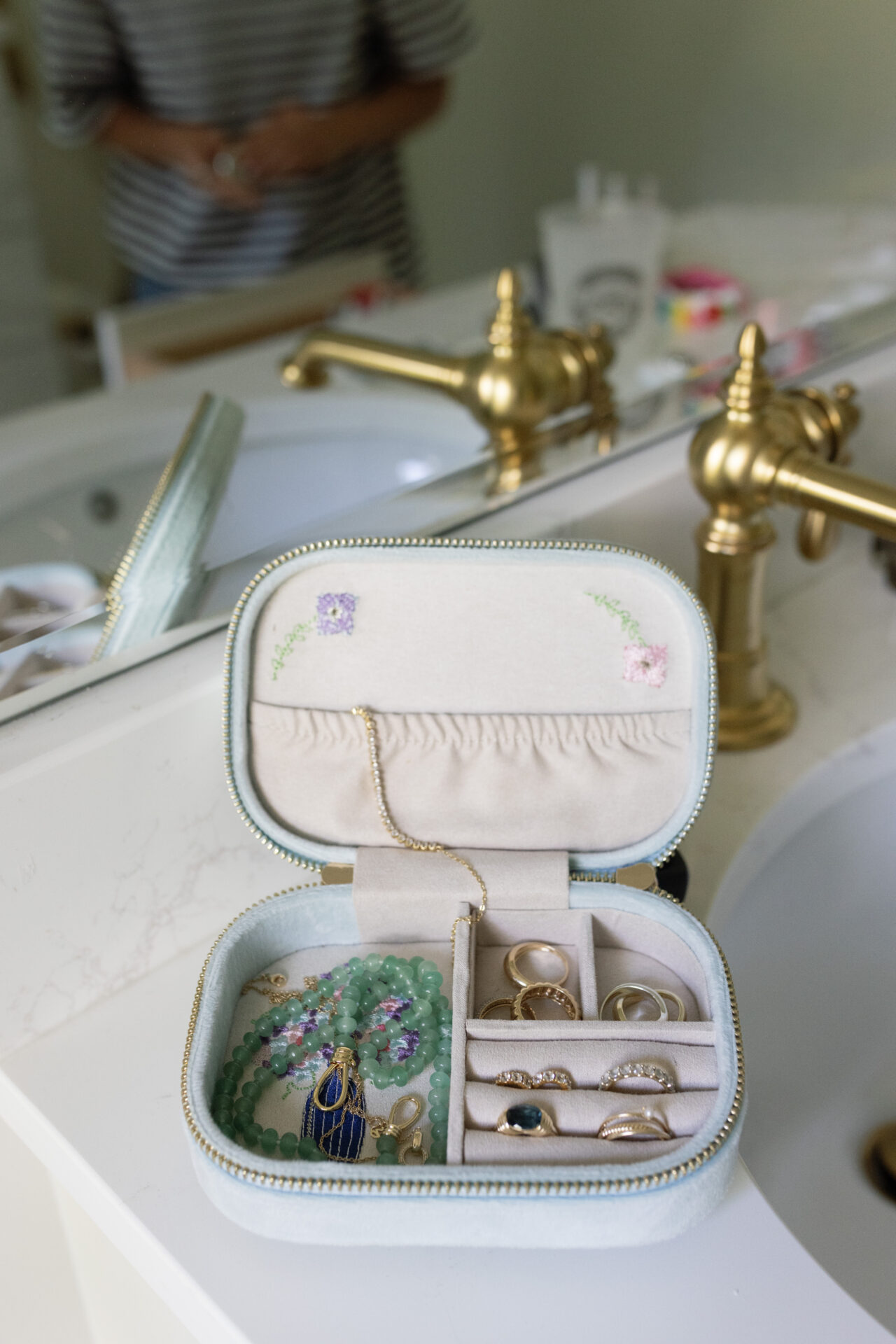 Open travel jewelry box filled with gold rings, a green bead bracelet, and a delicate gold necklace, sitting on a white marble bathroom counter with gold faucets