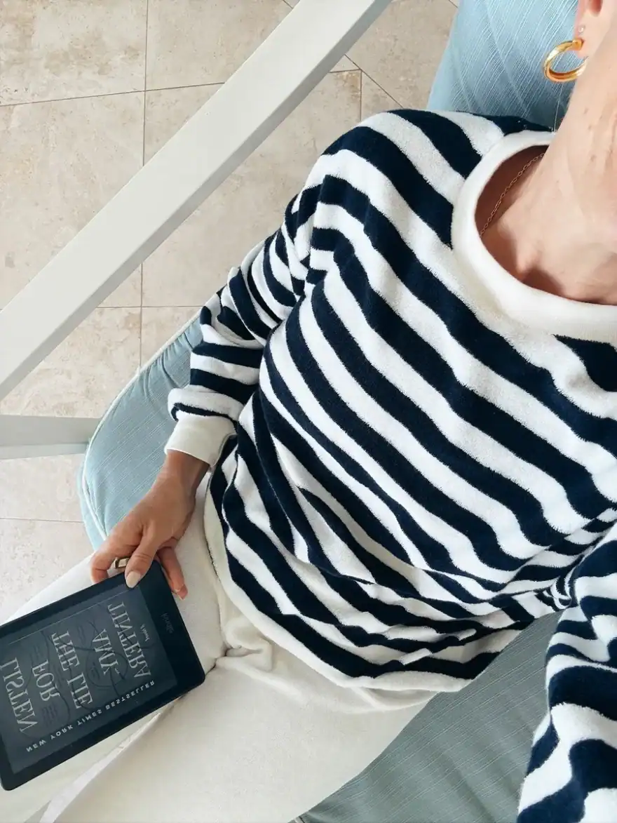 Woman sitting in chair reading Kindle wearing white pants and white and blue striped shirt