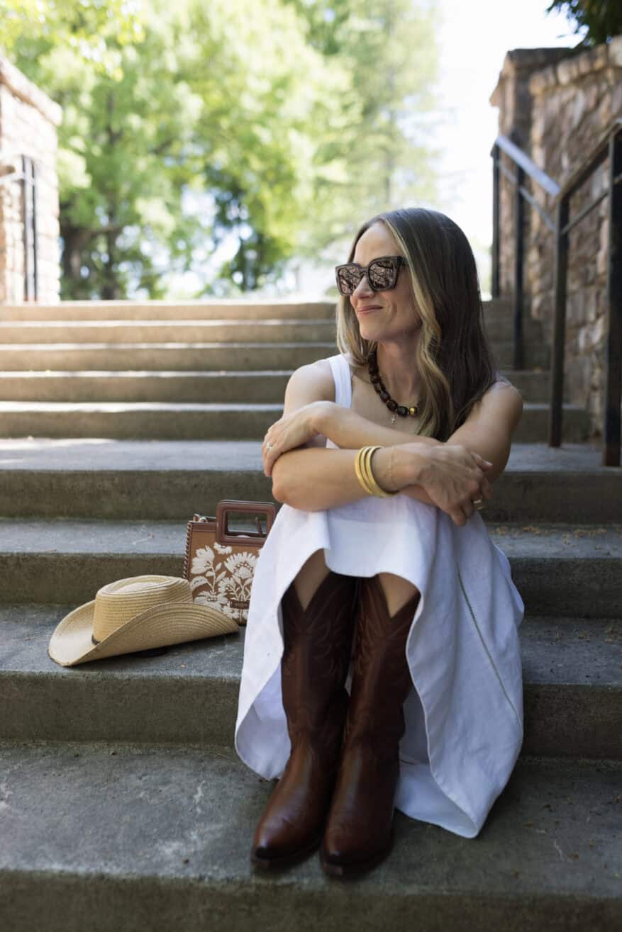 woman sitting on steps in leather cowboy boots and white linen dress with a cowboy hat