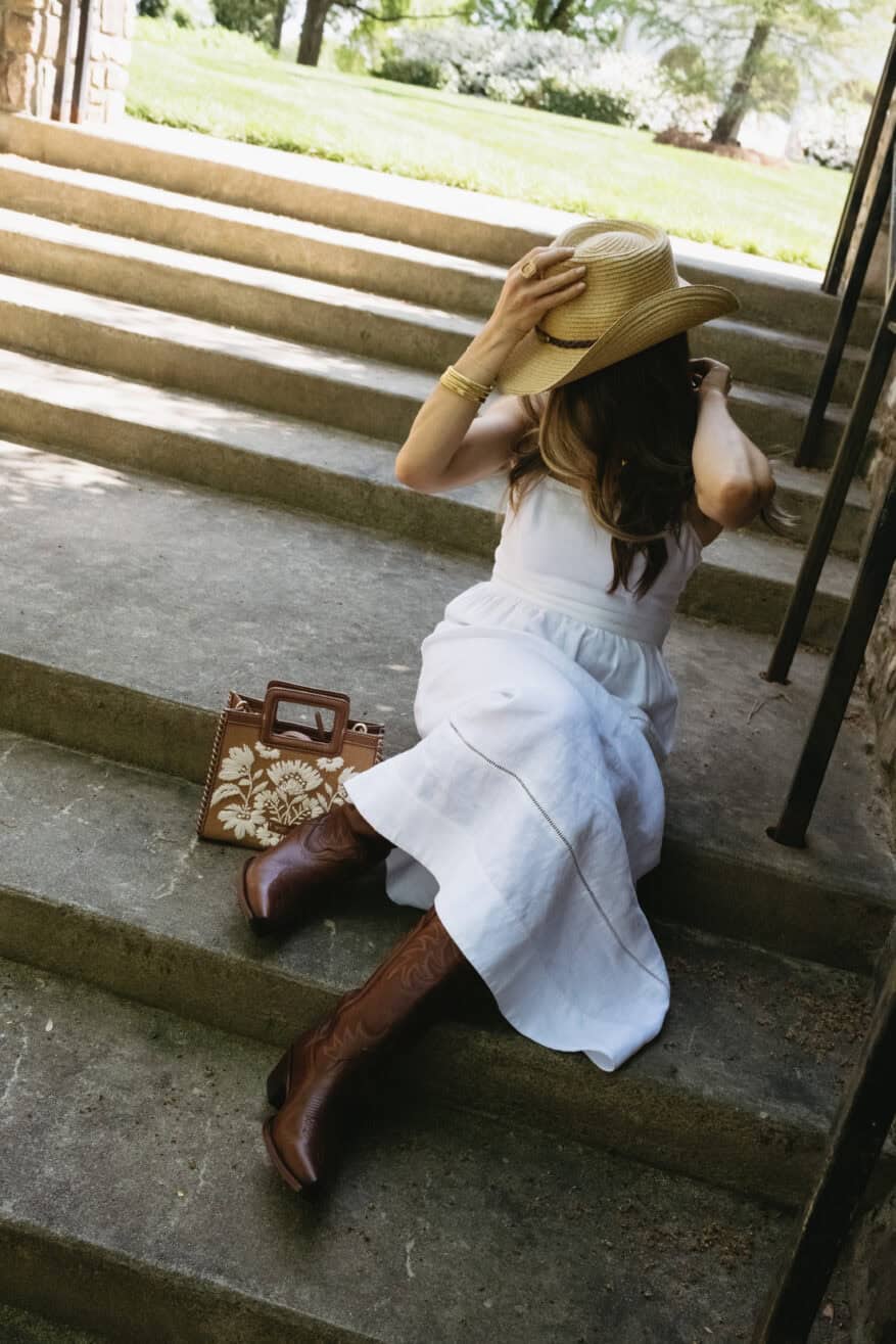 woman sitting on steps in leather cowboy boots and white linen dress with a cowboy hat on her head