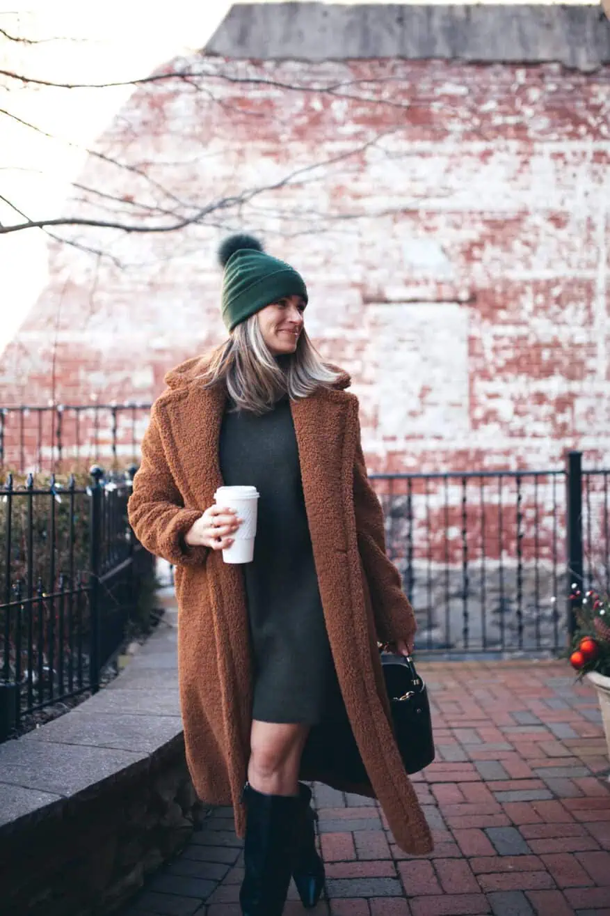 Woman walking while wearing a sweater dress with a long wool coat, boots, and a toboggan