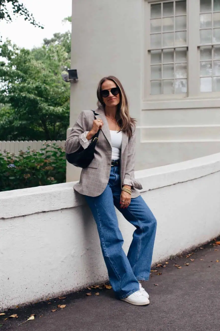 Woman leaning on wall wearing jeans, white top, and a fitted blazer with white sneakers