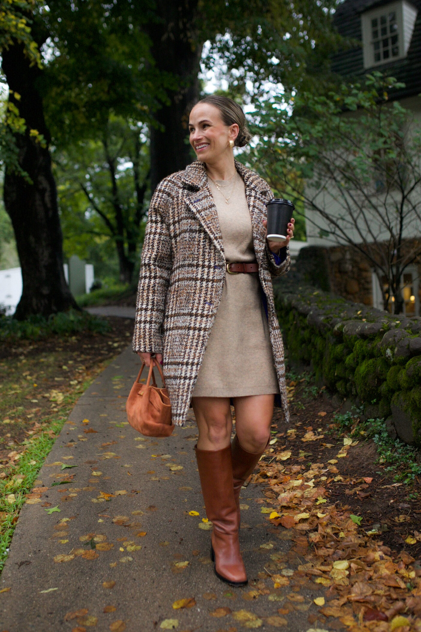 Woman walking in a sweater dress with a belt, tall boots, and a pea coat