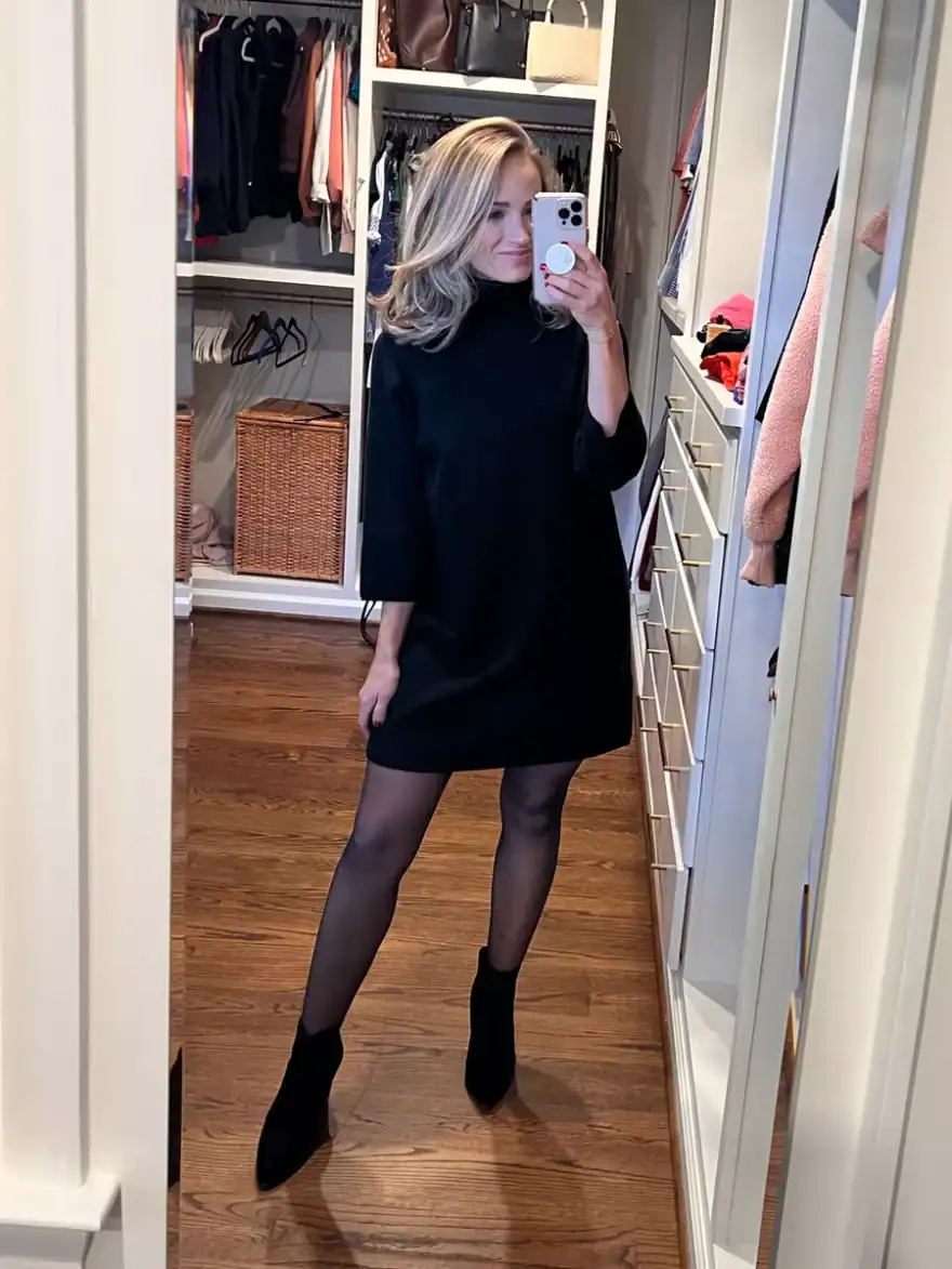 Woman taking a selfie while wearing a black sweater dress, opaque leggings, and booties
