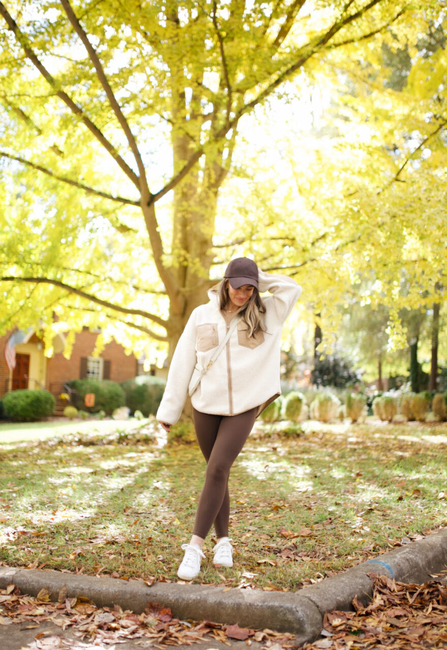 Woman walking in leaves wearing brown leggings and hat, cream jacket, and white shoes athleisure for women over 30