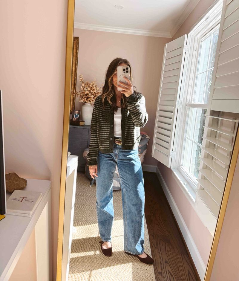 Woman taking a selfie with a white top under a cardigan, jeans, and a belt