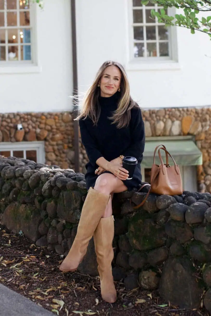 Woman sitting on rock wall wearing a black turtleneck sweater dress and tall boots