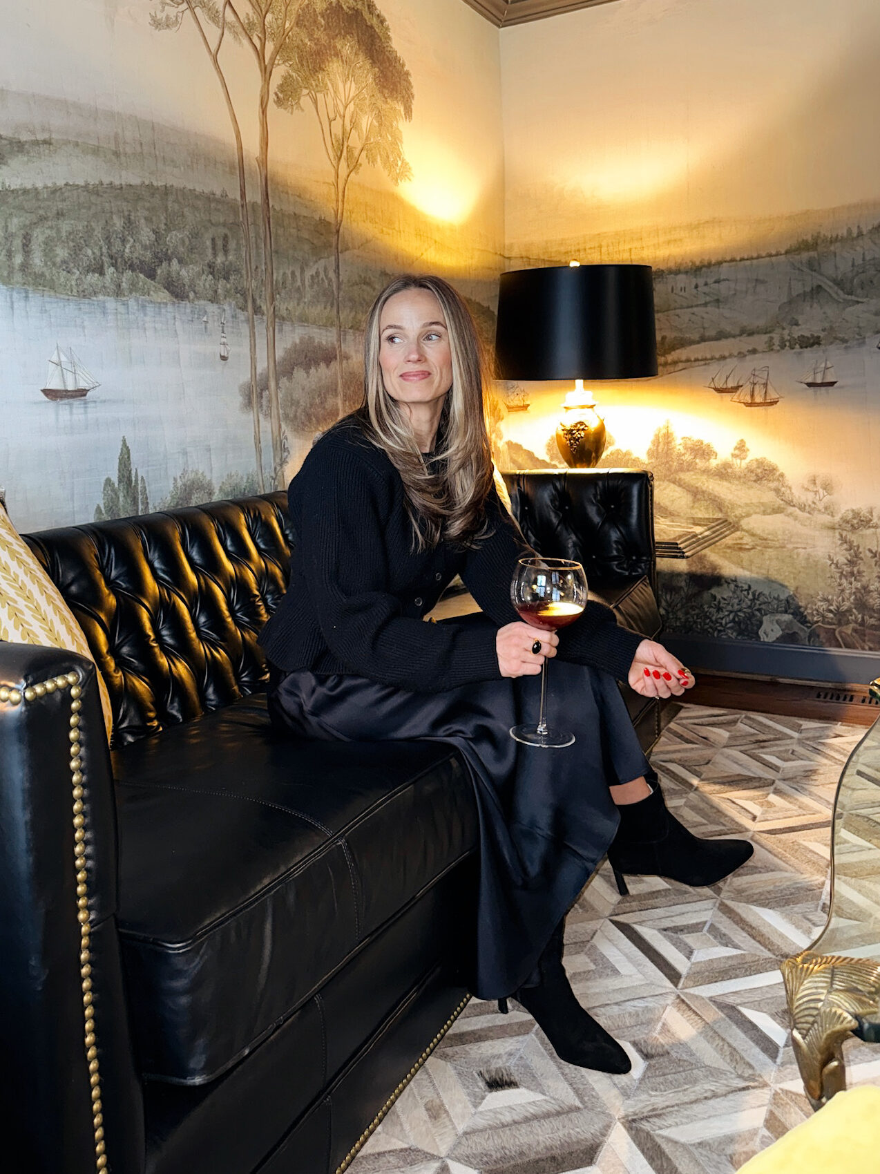 Woman sitting on couch with a glass of wine while wearing a black satin skirt, black cardigan, and black booties