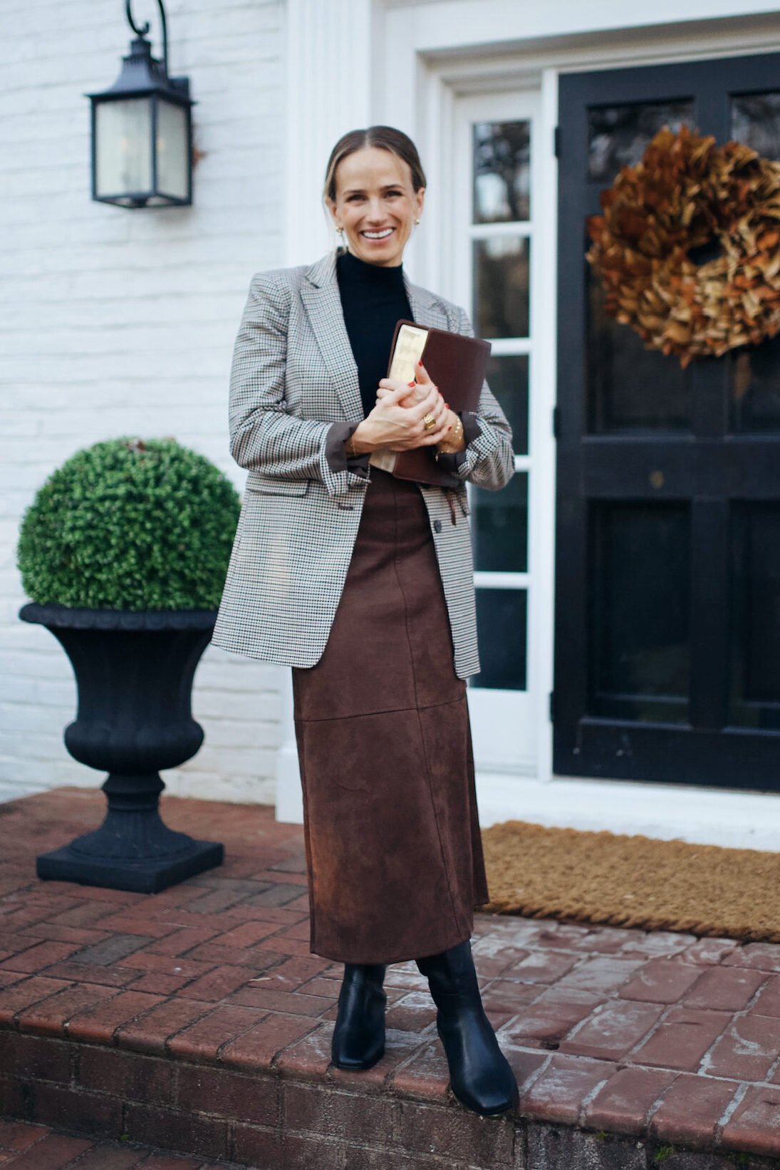Woman standing on porch waring suede skirt, knee high boots, and a blazer over a mock turtleneck