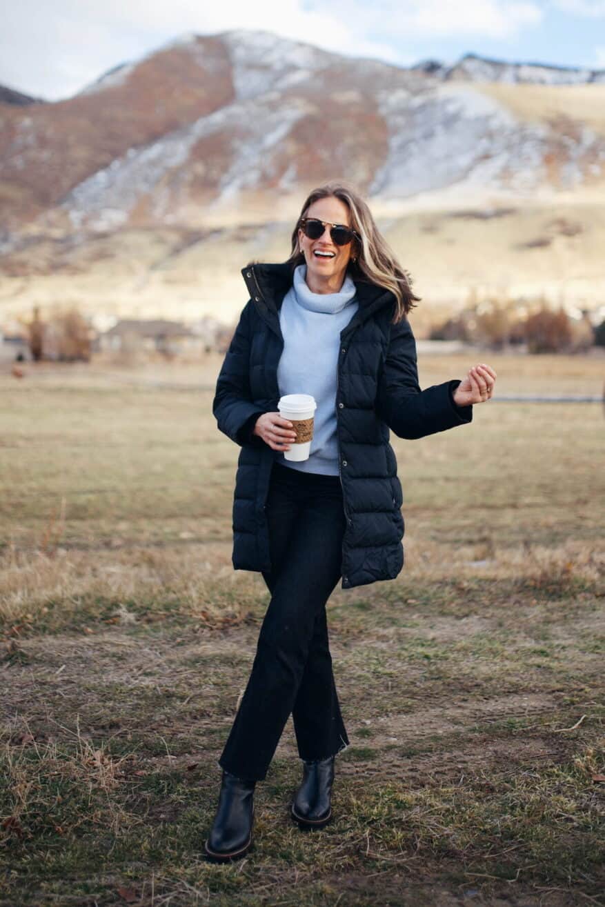 Woman standing in field with mountains in the background wearing black jeans, a blue turtleneck, and a puffer jacket