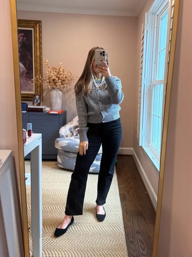 Woman taking a selfie while wearing black denim, ballet flats, and a button-up gray sweater