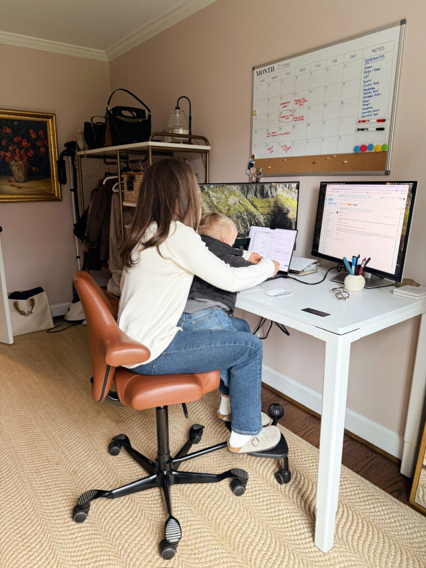 TeriLyn Adams wearing a cream sweater and jeans working at her desk while holding her baby