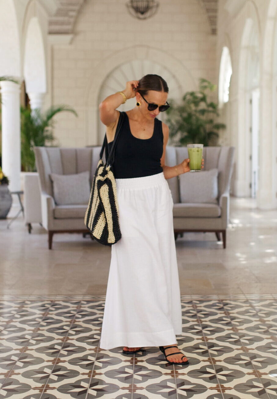 Woman standing in lobby with white skirt, black top, striped bag and sandals