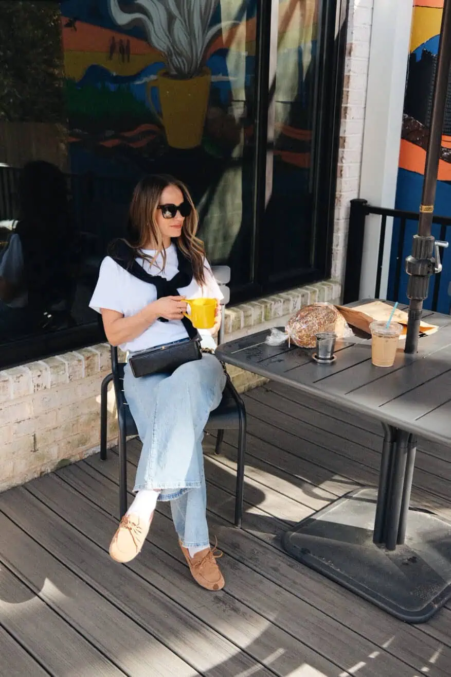 Woman sitting and having coffee wearing jeans, boat shoes, ruffle socks, white short sleeve top, and black sweater around shoulders
