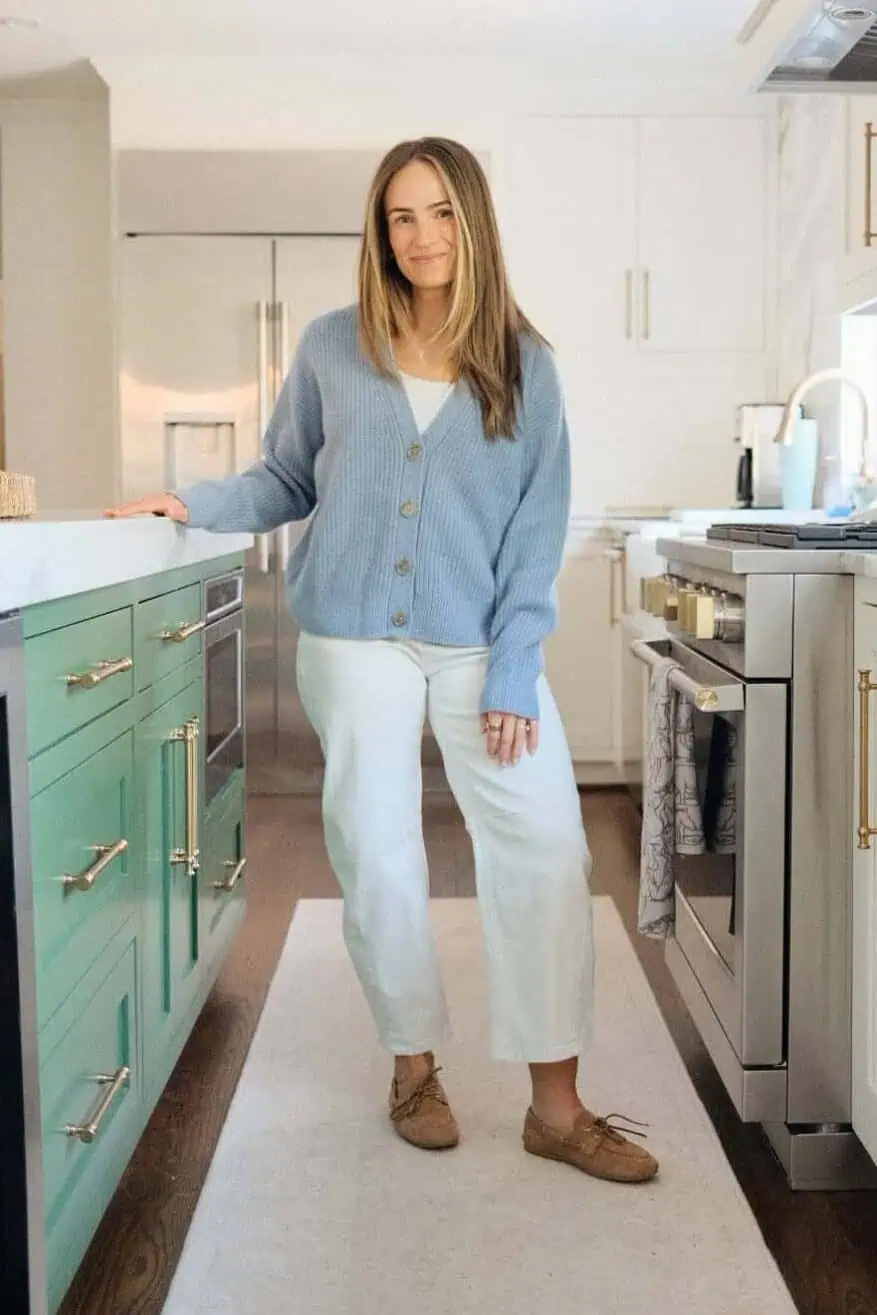 Woman wearing Quince blue cardigan with white jeans and loafer while standing in kitchen