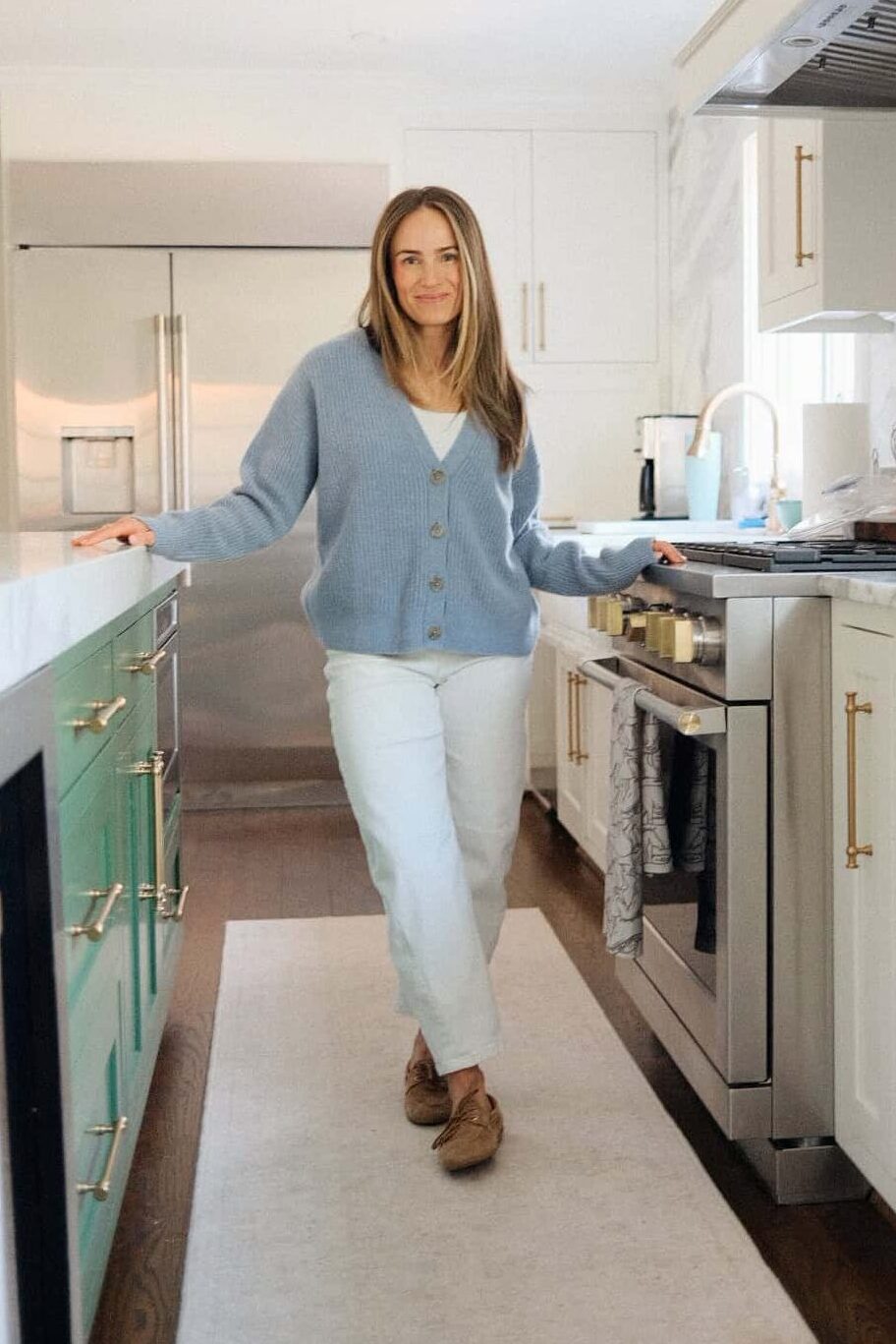 Woman wearing Quince blue cardigan with white top underneath with white jeans and loafer while standing in kitchen