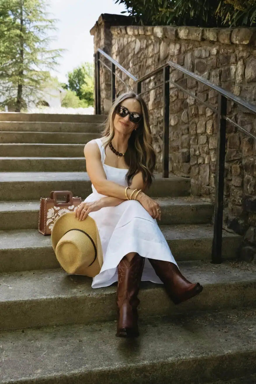 terilyn adams wearing white linen mid dress with cowboy boots and holding a bag sitting on the cement steps