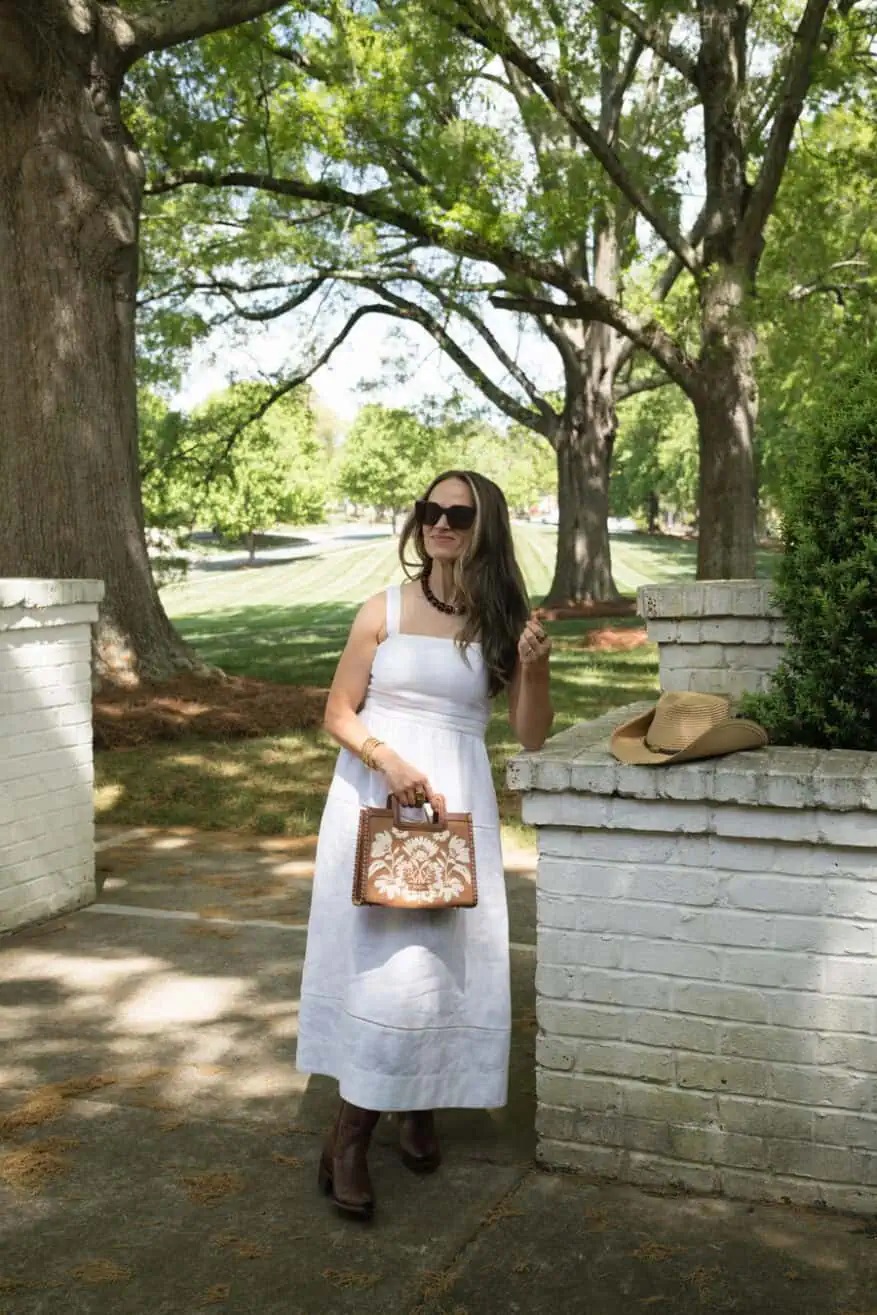 terilyn adams wearing white linen mid dress with cowboy boots and holding a bag in the park