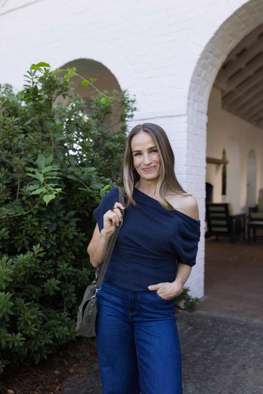 TeriLyn Adams wearing a navy Reformation one-shoulder structured top with dark wide-leg jeans and a suede crossbody